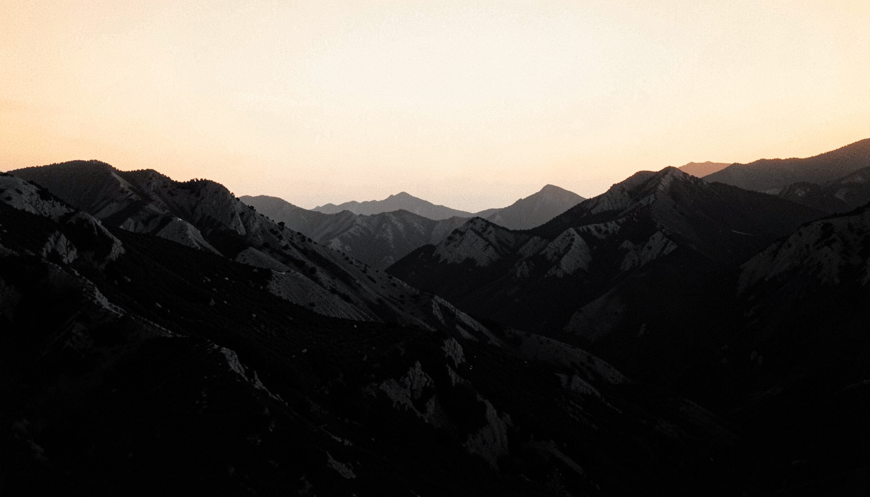 Dark atmospheric duotone photograph of rugged terrain at dusk, deep charcoal shadows and muted slate-gray ridges beneath a pale cream horizon.