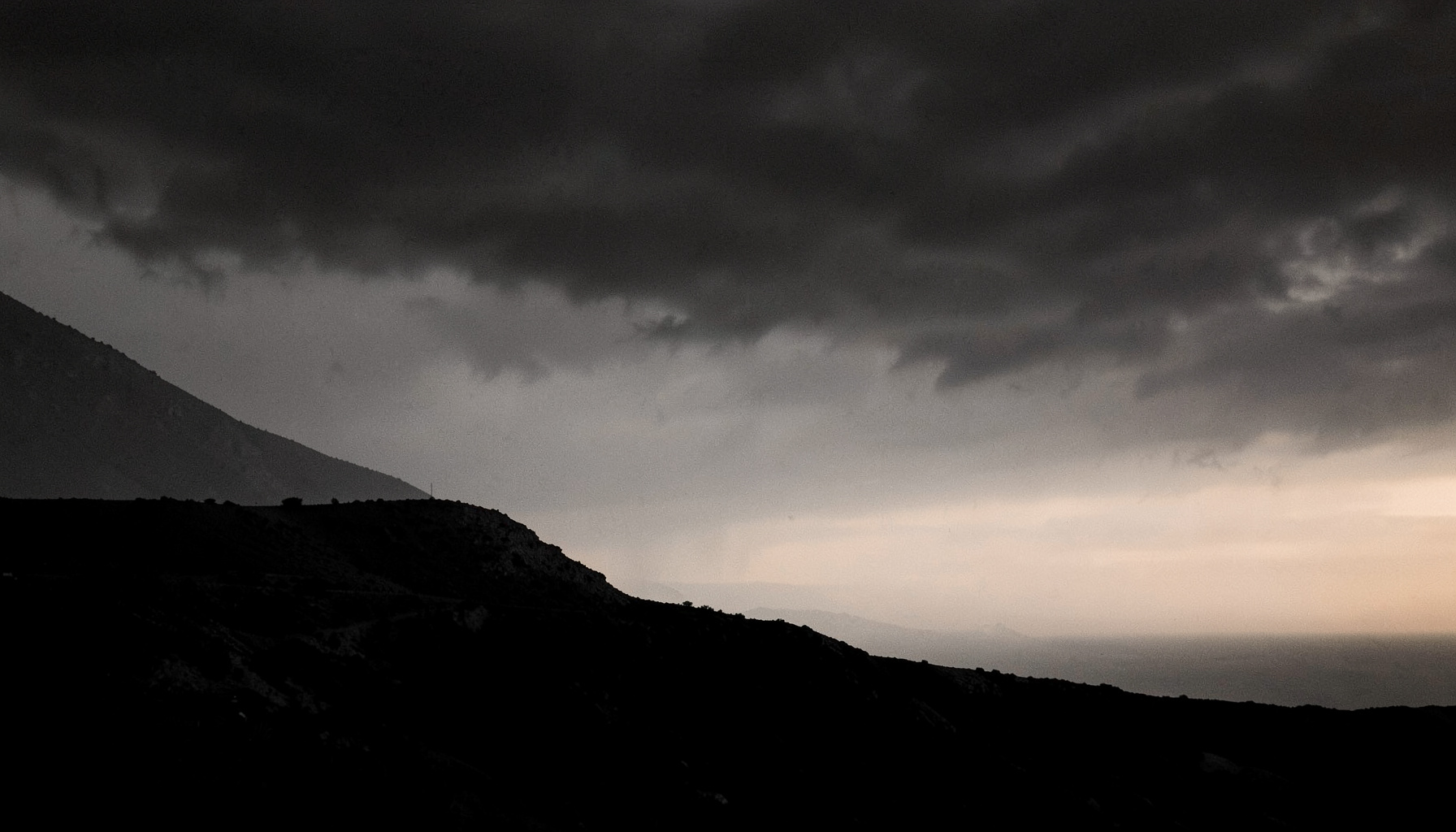 Wide duotone photograph of a stark ridgeline under a heavy sky, near-black charcoal tones meeting muted slate grays with a faint cream glow at the horizon.